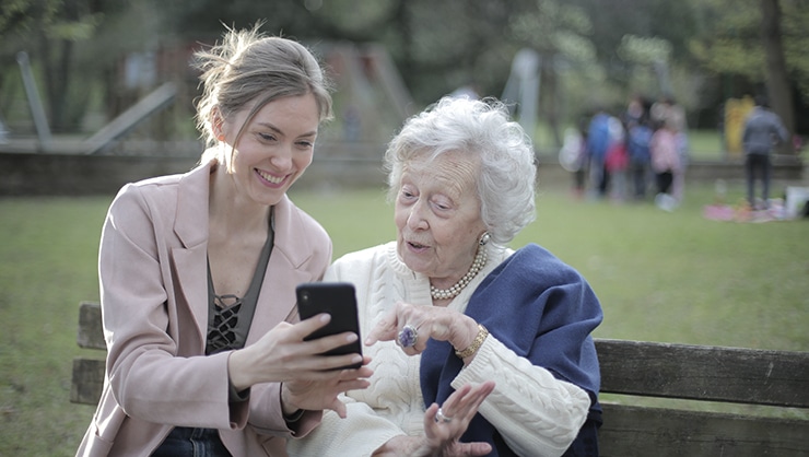 A smiling young blonde lady shows her smart phone to a fascinated white-haired woman