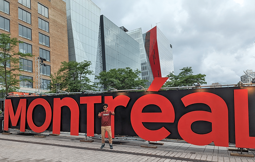 Young man poses in front of Montreal sign, giving two thumbs up.