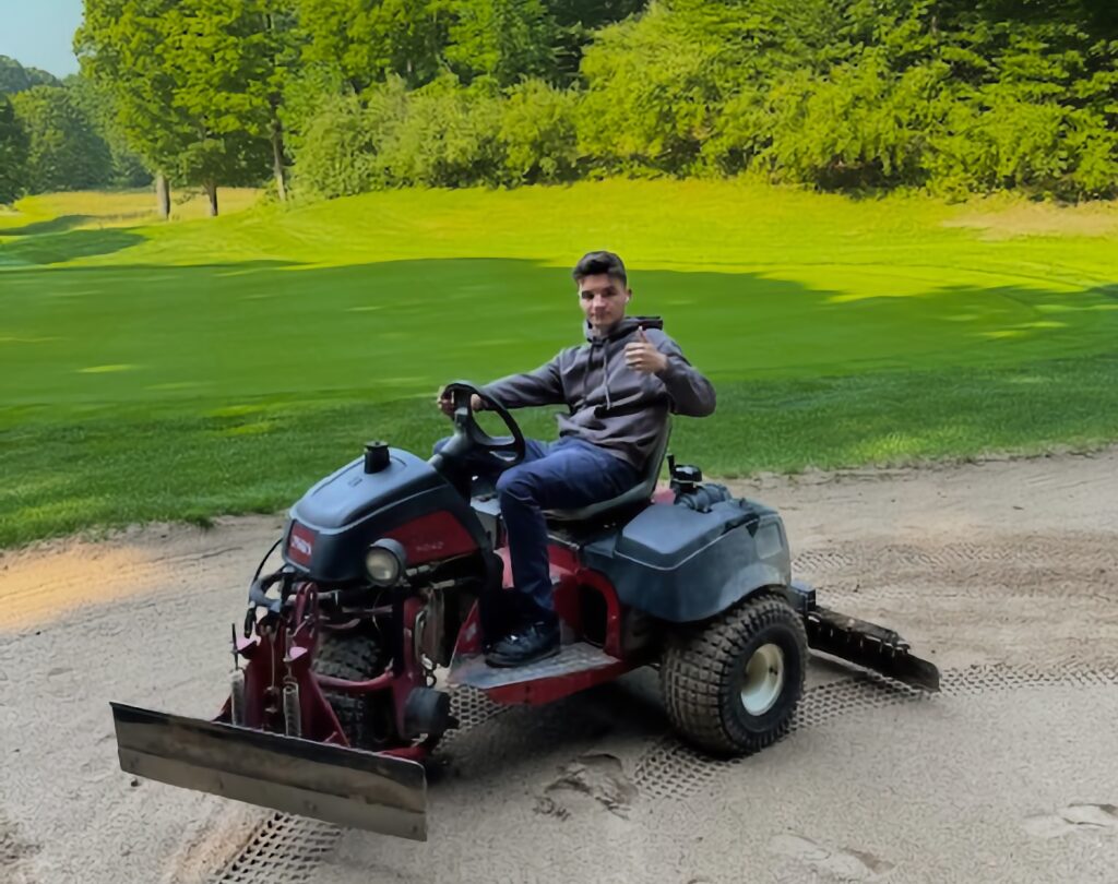 Young man sitting on a sand trap rake on a golf course.